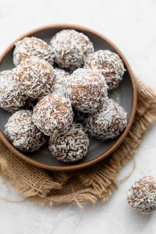 Date Coconut Rolls in a bowl on a burlap placemat. One outside of the bowl. Date rolls dusted in coconut.